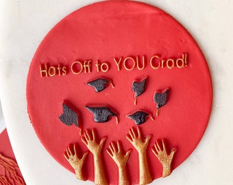May include: A close-up of graduation-themed decorations. A rectangular gray piece with the word "Congratulations!" in gold lettering. A red circle with the text "Hats Off to YOU Grad!" and graduation caps. A red rectangle with graduation caps.