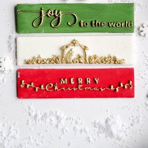 Op de afbeelding: Drie rode, witte en groene kerstfondanttoppers met gouden letters. De bovenste topper zegt "joy to the world", de middelste topper heeft een kerststal en de onderste topper zegt "merry christmas".