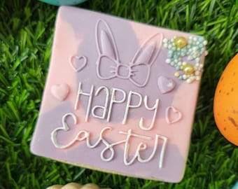May include: A pink and purple decorated sugar cookie with white icing that says "Happy Easter" and a bunny ear design. The cookie is surrounded by green grass.