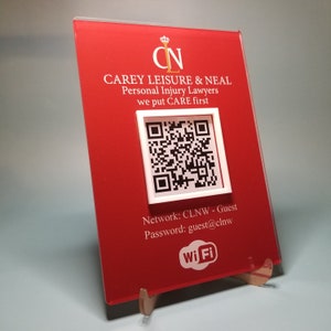 Może przedstawiać: Czerwony akrylowy znak z białym tekstem i kodem QR. Na znaku widnieje napis "CAREY LEISURE & NEAL Personal Injury Lawyers" i zawiera informacje o sieci Wi-Fi. Znak jest podparty przezroczystym stojakiem.