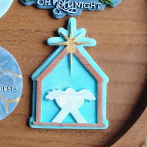 Op de afbeelding: Vier blauwe en gouden kerstversieringen met religieuze thema's. De ornamenten bevatten de tekst "Oh Holy Night", "O Come Let Us Adore Him", en "A Child Has Been Born For Us, A Son Is Given To Us: Authority Rests Upon Him, And He Is Named Jesus."