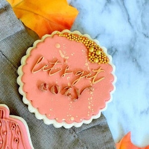 Op de afbeelding: Drie roze koekjes met witte geschulpte randen. Twee koekjes hebben gouden tekst met de tekst "sweater weather" en "let's get cozy". Het derde koekje heeft de vorm van een trui. Gouden sprinkles sieren de koekjes.
