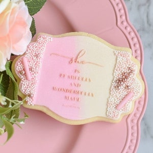 May include: A pink and white decorated sugar cookie with the text "She is fearless and a wonderful wife" in gold lettering.