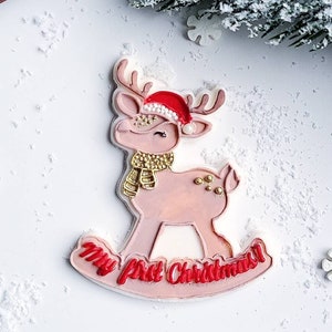 Op de afbeelding: Een roze en witte rendierkoekje met gouden glitter en een rode kerstmanmuts. Het koekje is versierd met de woorden "My First Christmas!" in rode glazuur.