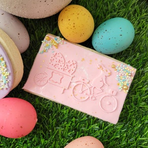 May include: Pink and white decorated sugar cookie with a bunny riding a bicycle and carrying a basket of eggs. The cookie is surrounded by pastel colored speckled Easter eggs on a green grass background.