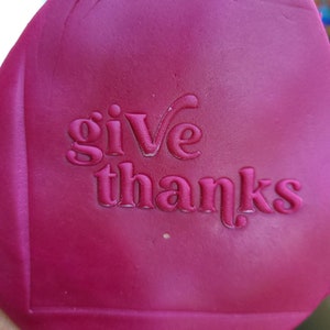 Op de afbeelding: Een close-up van een roze rubberen stempel met de woorden "give thanks" erop gegraveerd.
