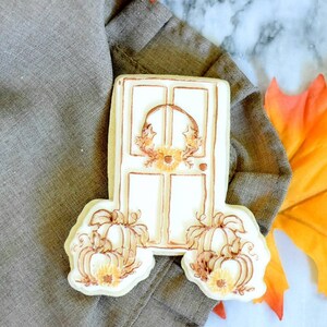 Op de afbeelding: Een witte suikerkoek versierd met bruine glazuur om eruit te zien als een deur met een krans en pompoenen. De koek ligt op een bruine stoffen servet met een herfstblad.