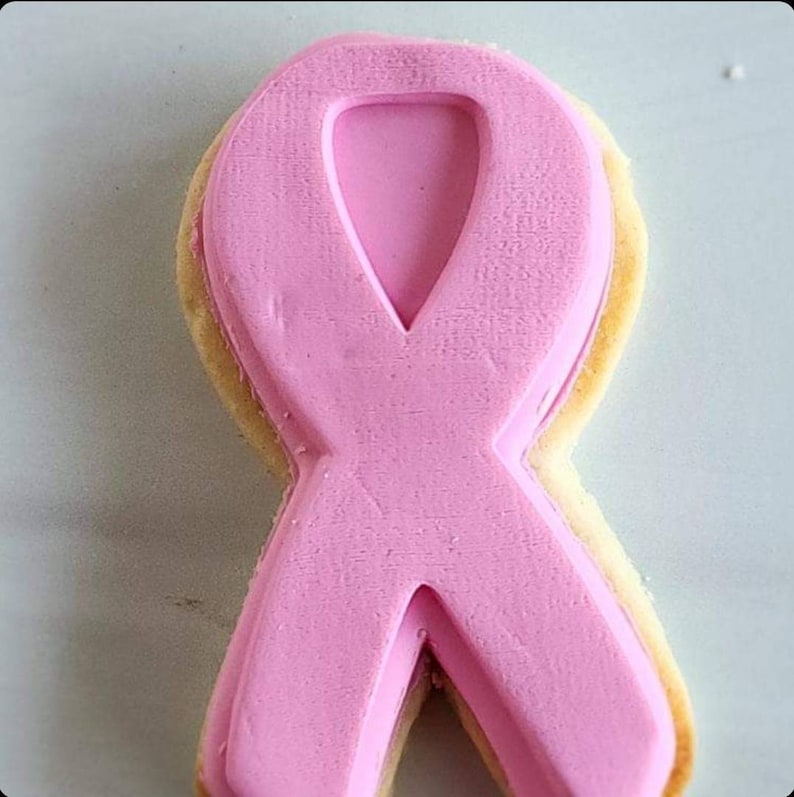 Pu&ograve; includere: Biscotto a forma di nastro rosa decorato con glassa rosa. Il biscotto ha la forma di un nastro e presenta un design in rilievo.