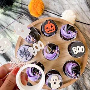 Op de afbeelding: Een houten dienblad met negen Halloween-thema cupcakes. De cupcakes zijn versierd met paarse en zwarte frosting en bedekt met verschillende Halloween-decoraties, waaronder geesten, pompoenen, spinnen en het woord "BOO".
