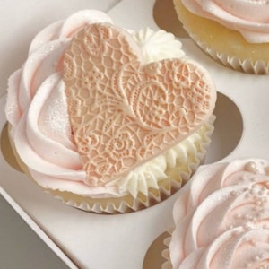 May include: Three pink and white cupcakes with delicate frosting designs. One cupcake features a heart-shaped lace design.