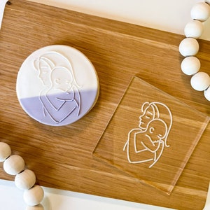 May include: A white and purple cookie with a line drawing of a mother and child hugging. The cookie is on a wooden surface next to a clear acrylic stamp with the same design.