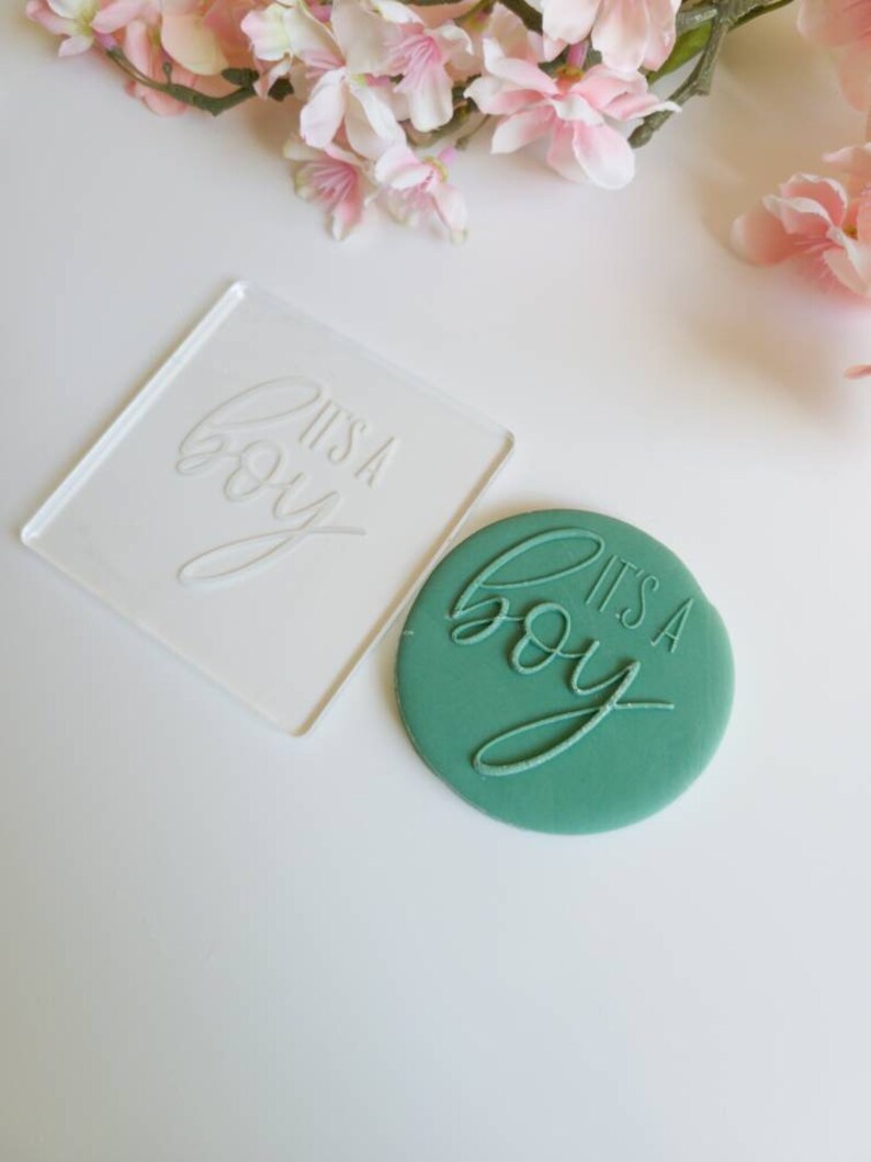 Op de afbeelding: Een transparante acryl stempel met de tekst "It's a boy" en een groene fondantkoekje met dezelfde tekst in reli&euml;f.
