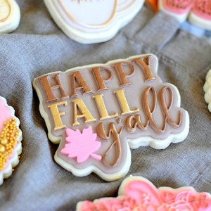 Op de afbeelding: Een close-up van een gedecoreerde suikerkoek met de woorden "Happy Fall y'all" in gouden en bruine glazuur. De koek is versierd met een roze blad en wordt omringd door andere gedecoreerde koekjes.