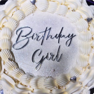 Puede incluir: Un pastel blanco con glaseado blanco y un acabado de purpurina plateada. El pastel está decorado con corazones plateados y las palabras "Birthday Girl" en letras plateadas.