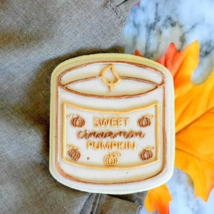 Op de afbeelding: Een witte suikerkoek versierd met bruine en oranje glazuur. De koek is gevormd als een kaarsglas en heeft het opschrift "Sweet Cinnamon Pumpkin" op het etiket.