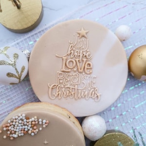 May include: Two round, beige cookies decorated with edible icing. One cookie has a Christmas tree design with the words "Joy, Hope, Love, Peace, Christmas". The other cookie has a nativity scene design with a star, palm trees, and three camels.