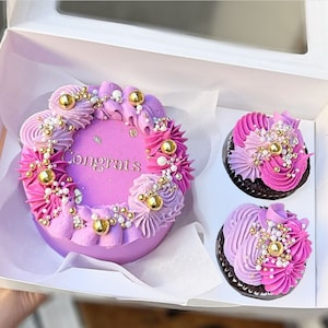Könnte beinhalten: Ein lila und rosa Kuchen mit goldenen Streuseln und dem Wort "Congrats" in Gold auf der Oberseite. Der Kuchen befindet sich in einer weißen Schachtel mit zwei Cupcakes daneben.