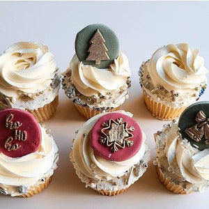 Op de afbeelding: Zes gedecoreerde cupcakes met verschillende kerstthema's. De cupcakes zijn bedekt met witte frosting en versierd met groene, gouden en rode accenten. Sommige van de decoraties omvatten een kerstboom, een sneeuwvlok en een bel.