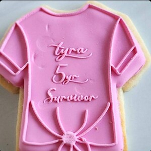 Pu&ograve; includere: Biscotto di zucchero rosa decorato con glassa rosa a forma di maglietta. La glassa ha le parole "Tyra 5yr Survivor" scritte su di essa.