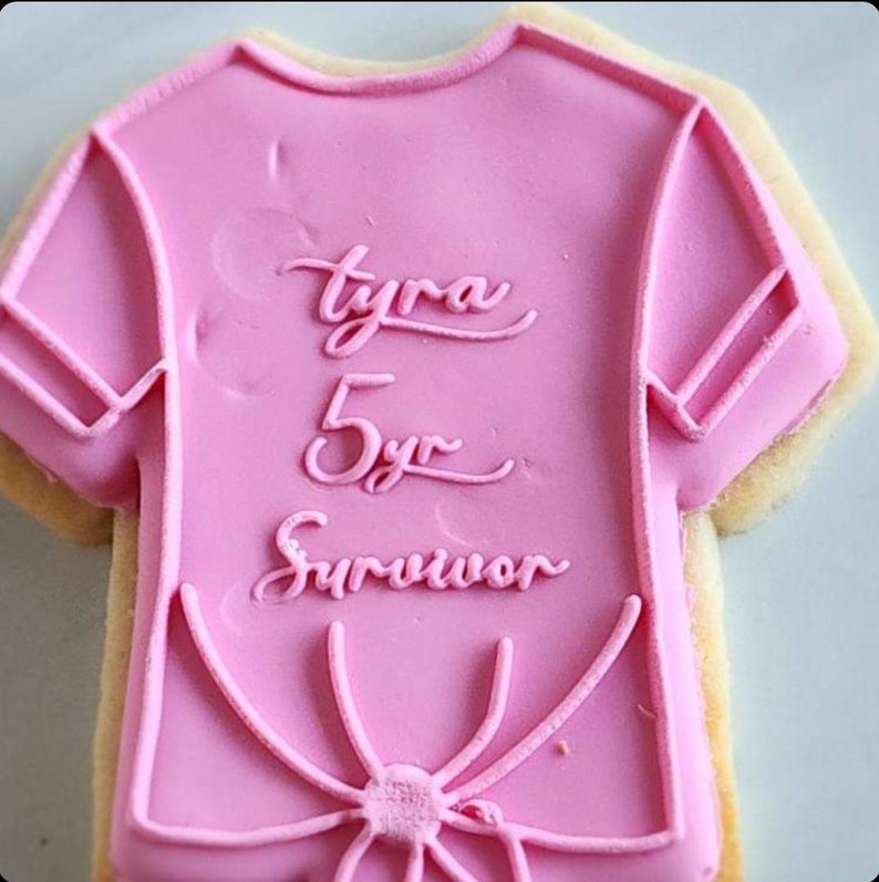 Pu&ograve; includere: Biscotto di zucchero rosa decorato con glassa rosa a forma di maglietta. La glassa ha le parole "Tyra 5yr Survivor" scritte su di essa.