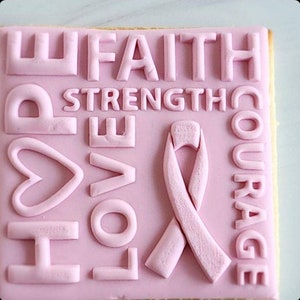 Pu&ograve; includere: Biscotto di zucchero rosa decorato con glassa rosa e in rilievo con le parole "HOPE", "LOVE", "FAITH", "STRENGTH", "COURAGE" e un nastro rosa.