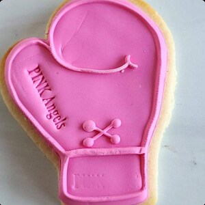 Pu&ograve; includere: Biscotto di zucchero rosa decorato con glassa rosa a forma di guanto da boxe. La glassa ha le parole "PINK Angels" scritte su di essa.