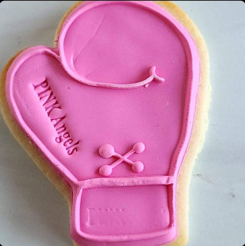 Pu&ograve; includere: Biscotto di zucchero rosa decorato con glassa rosa a forma di guanto da boxe. La glassa ha le parole "PINK Angels" scritte su di essa.