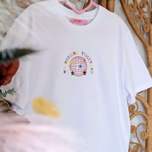 May include: White t-shirt with the embroidered phrase "WE BELONG TOGETHER" surrounding a globe design with colorful stars. The shirt is hanging on a pink heart-shaped hanger against a woven background.