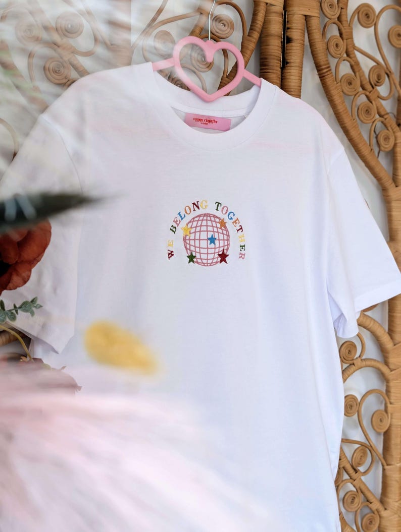 May include: White t-shirt with the embroidered phrase "WE BELONG TOGETHER" surrounding a globe design with colorful stars. The shirt is hanging on a pink heart-shaped hanger against a woven background.