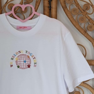 May include: A white t-shirt with the embroidered phrase "WE BELONG TOGETHER" encircling a globe design with colorful stars. The shirt is hanging on a pink heart-shaped hanger against a woven background.
