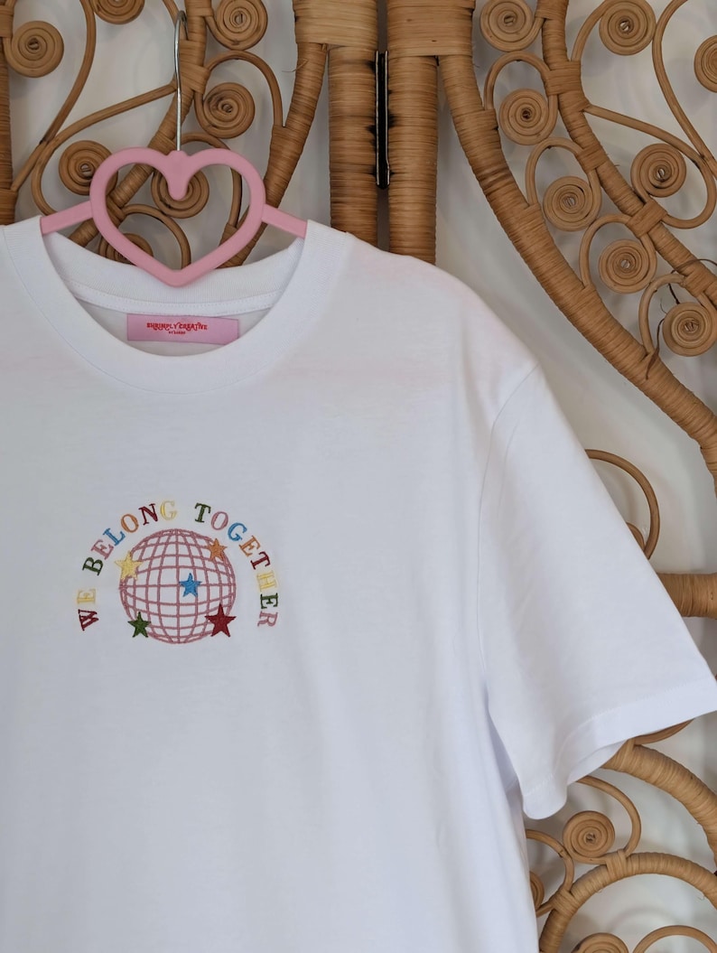 May include: A white t-shirt with the embroidered phrase "WE BELONG TOGETHER" encircling a globe design with colorful stars. The shirt is hanging on a pink heart-shaped hanger against a woven background.