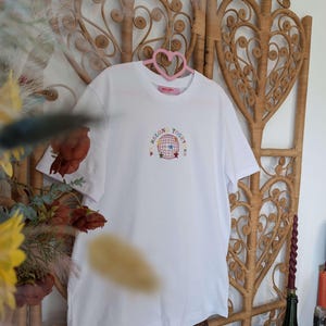 May include: White t-shirt with the embroidered text "WE BELONG TOGET[HER]" around a globe design with stars. The shirt is hanging on a pink heart-shaped hanger against a woven wicker backdrop. The shirt is a casual wear item.