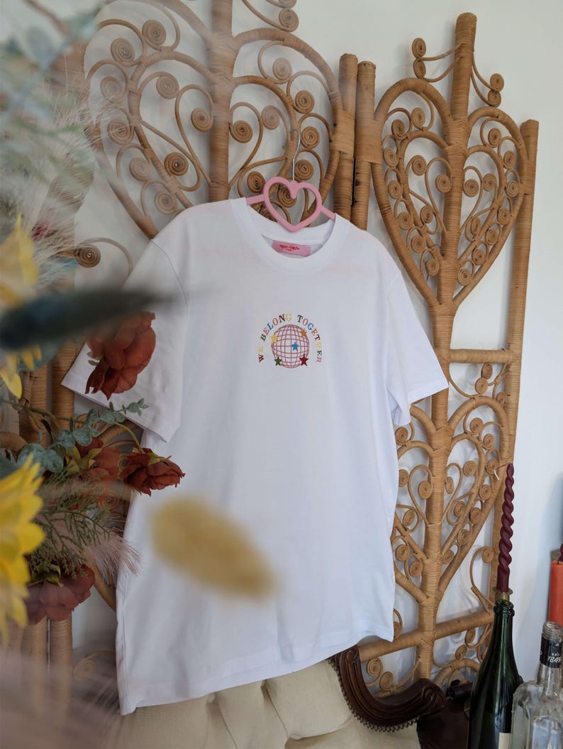 May include: White t-shirt with the embroidered text "WE BELONG TOGET[HER]" around a globe design with stars. The shirt is hanging on a pink heart-shaped hanger against a woven wicker backdrop. The shirt is a casual wear item.