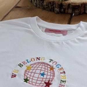 May include: White t-shirt with embroidered design. The design features a globe with a grid pattern, surrounded by the words "WE BELONG TOGETHER" in various colors. Colorful stars are also embroidered around the globe.
