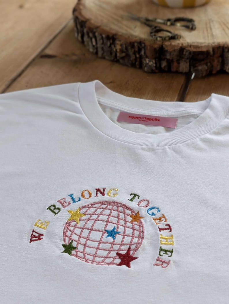 May include: White t-shirt with embroidered design. The design features a globe with a grid pattern, surrounded by the words "WE BELONG TOGETHER" in various colors. Colorful stars are also embroidered around the globe.