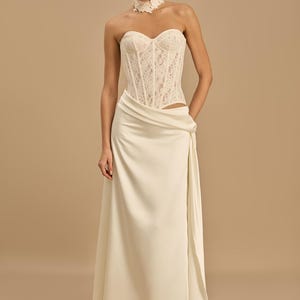 May include: An ivory wedding dress with a lace corset and draped skirt. The dress includes a matching lace choker. The style is elegant and suitable for bridal wear. The dress is floor length.