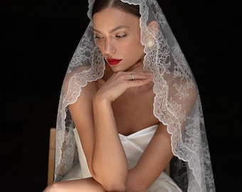 May include: A bride wearing a white wedding dress and a white lace veil. She is sitting with her arms crossed and looking down.
