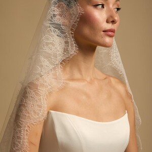 May include: A white bridal veil with intricate lace detailing along the edges. The veil is draped over a white strapless dress. The image is a side profile, showcasing the delicate texture and design of the veil.