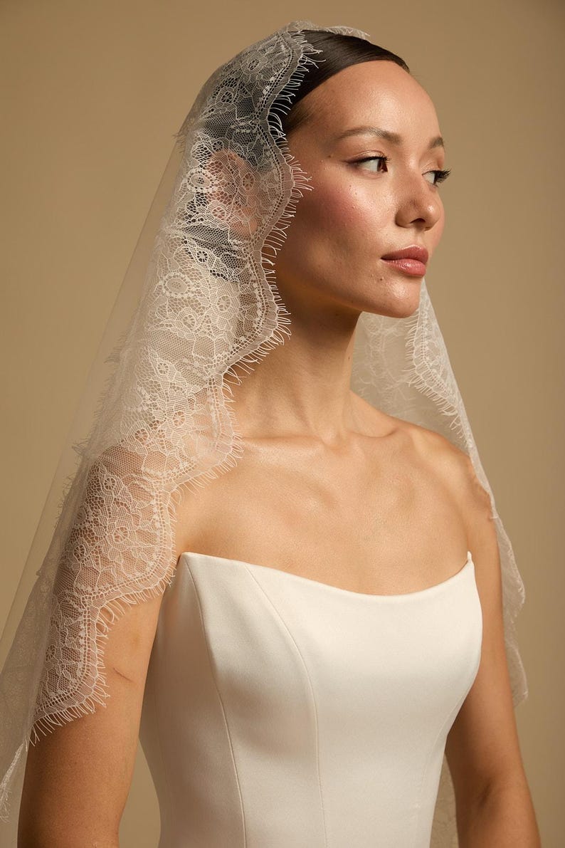 May include: A white bridal veil with intricate lace detailing along the edges. The veil is draped over a white strapless dress. The image is a side profile, showcasing the delicate texture and design of the veil.
