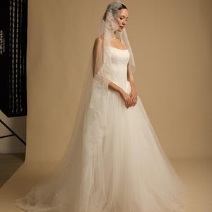 May include: A white wedding dress with a tulle skirt and a square neckline. The bride wears a veil with lace trim. The background is a neutral beige. The dress is elegant and classic.
