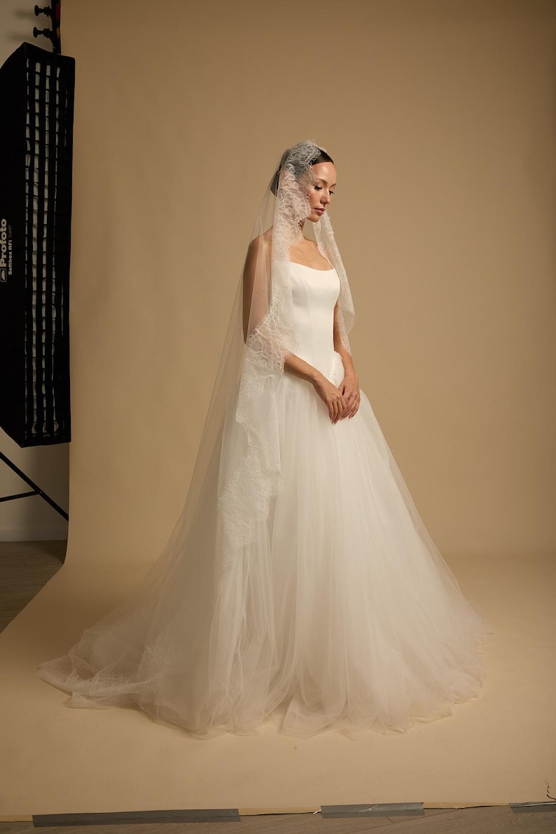 May include: A white wedding dress with a tulle skirt and a square neckline. The bride wears a veil with lace trim. The background is a neutral beige. The dress is elegant and classic.
