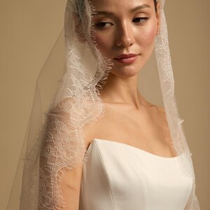 May include: A bride wearing a white strapless wedding dress and a sheer veil. The veil is trimmed with delicate lace detailing. The background is a soft, neutral color.