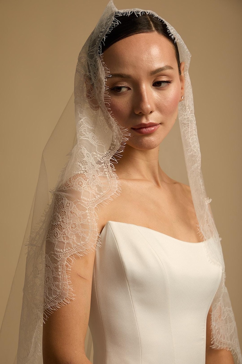 May include: A bride wearing a white strapless wedding dress and a sheer veil. The veil is trimmed with delicate lace detailing. The background is a soft, neutral color.