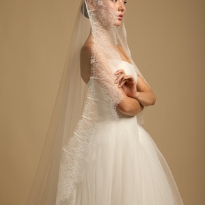 May include: A bride in a white wedding dress and veil. The veil is sheer with delicate lace trim along the edges. The dress is a strapless design with a full skirt. The background is a solid tan color.