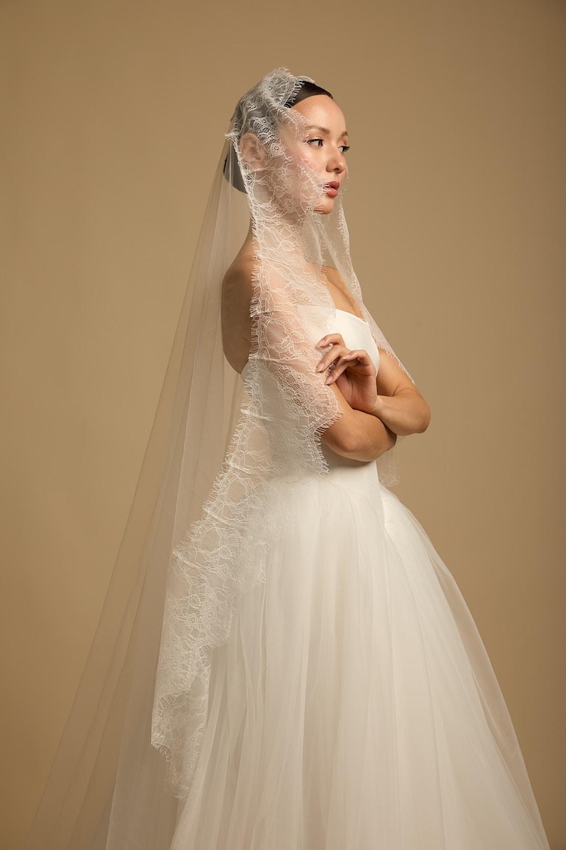 May include: A bride in a white wedding dress and veil. The veil is sheer with delicate lace trim along the edges. The dress is a strapless design with a full skirt. The background is a solid tan color.