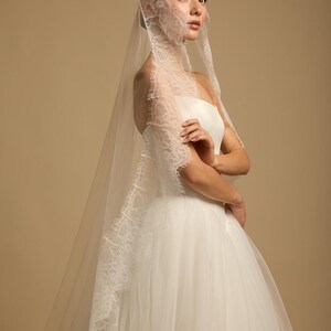 May include: A bride in a white strapless wedding dress and a long, sheer veil with delicate lace trim. The veil cascades down the back, and the bride is posed with her arms crossed, creating an elegant and romantic image.