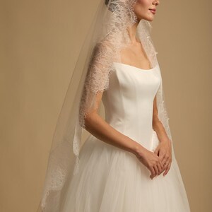 May include: A white wedding dress with a tulle skirt and a square neckline. The bride wears a delicate, sheer veil with intricate lace trim. The veil cascades down the back, adding a touch of elegance to the bridal ensemble.