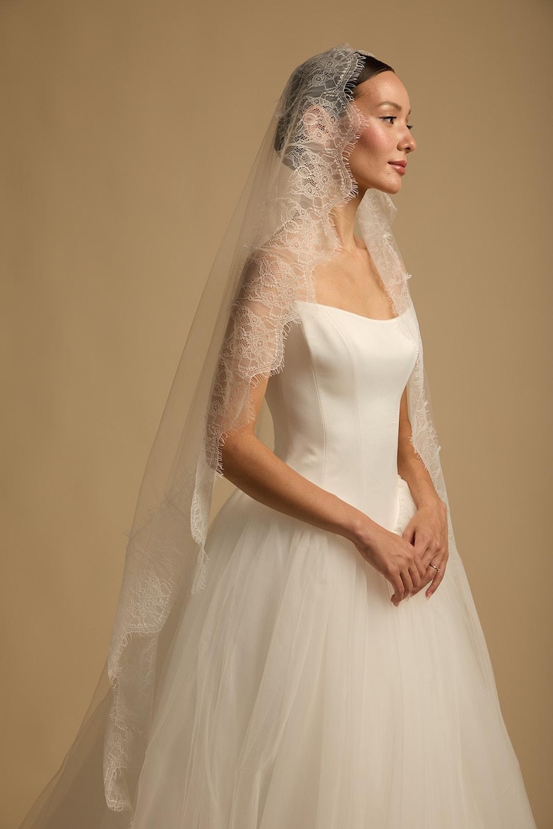 May include: A white wedding dress with a tulle skirt and a square neckline. The bride wears a delicate, sheer veil with intricate lace trim. The veil cascades down the back, adding a touch of elegance to the bridal ensemble.