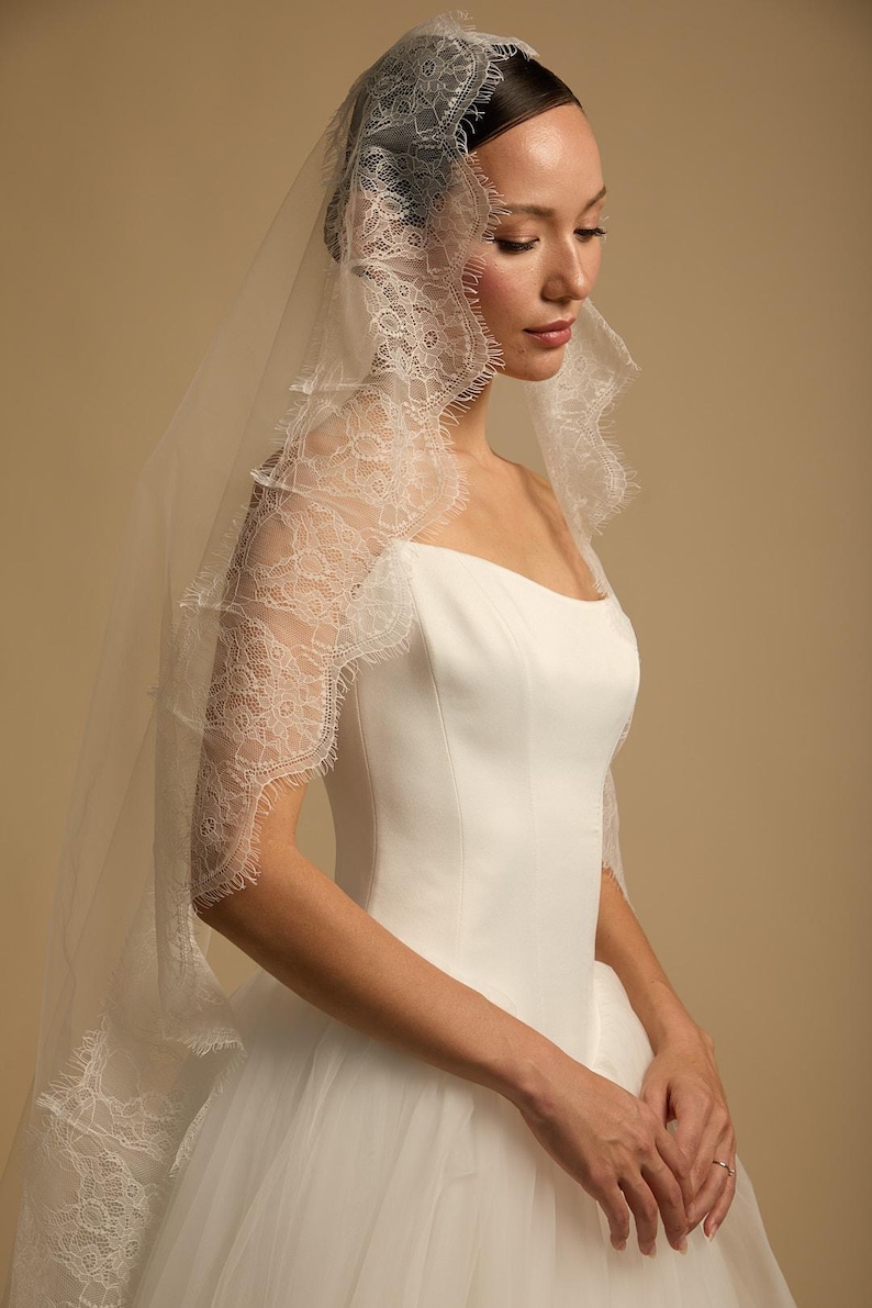 May include: A white wedding dress with a square neckline and a tulle skirt. The bride is wearing a delicate, sheer veil with intricate lace detailing along the edges. The veil drapes over the shoulders and frames the face.