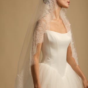 May include: A white wedding dress with a square neckline and a tulle skirt. The bride is wearing a delicate lace-trimmed veil. The veil is draped over the head and shoulders, creating a soft, romantic look. The dress is elegant and classic.